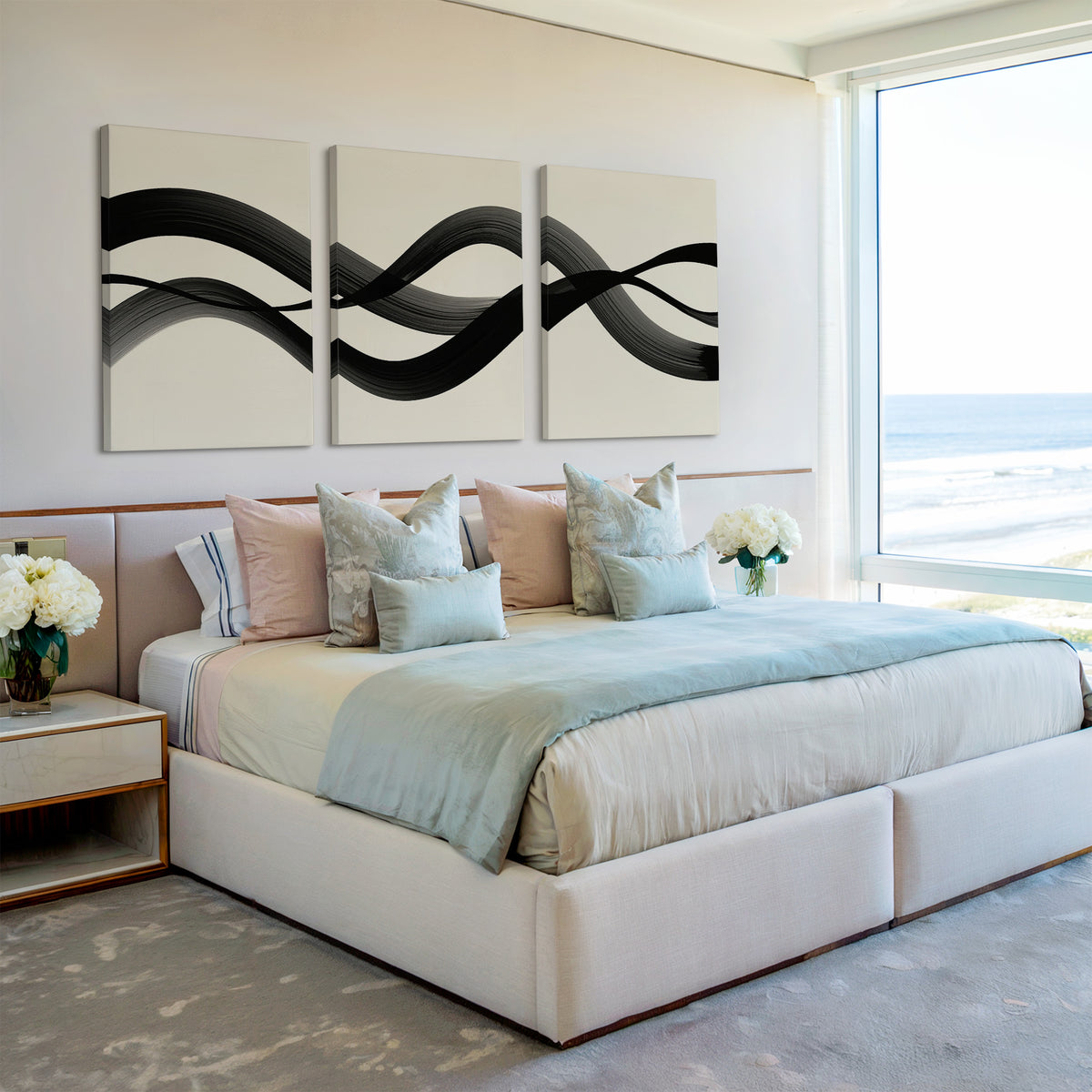 A modern bedroom in Nordic Park features a large bed with light blue and beige bedding, decorative pillows, two side tables with white flowers, a Fluid Curves Set of 3 wall art, and a large window offering an ocean view.