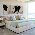 A modern bedroom features a large bed with pastel bedding, several pillows, and the Monochrome Flora Set of 3 botanical wall art above. Flower arrangements decorate the bedside tables, and a big window reveals an ocean view.