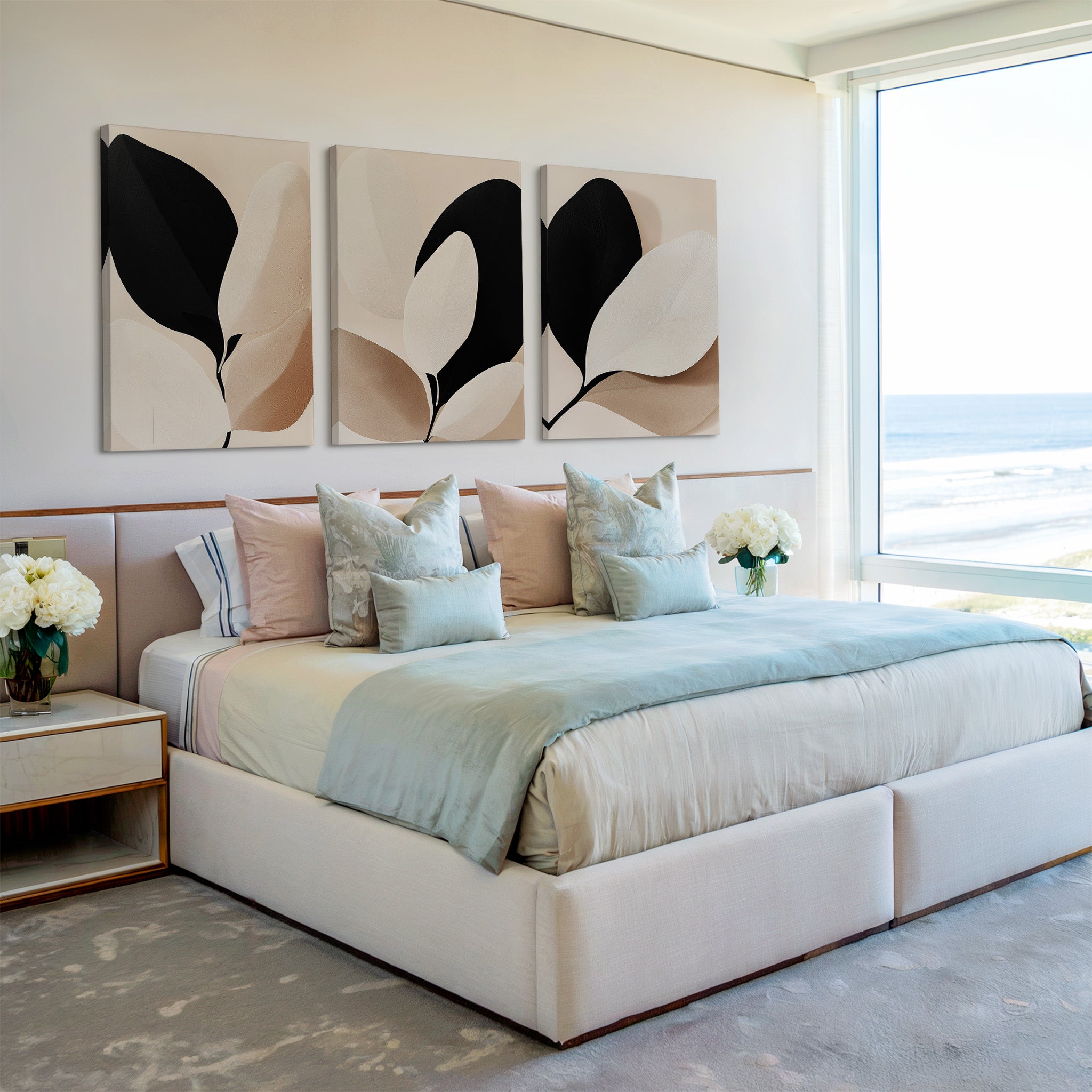 A modern bedroom features a large bed with pastel bedding, several pillows, and the Monochrome Flora Set of 3 botanical wall art above. Flower arrangements decorate the bedside tables, and a big window reveals an ocean view.