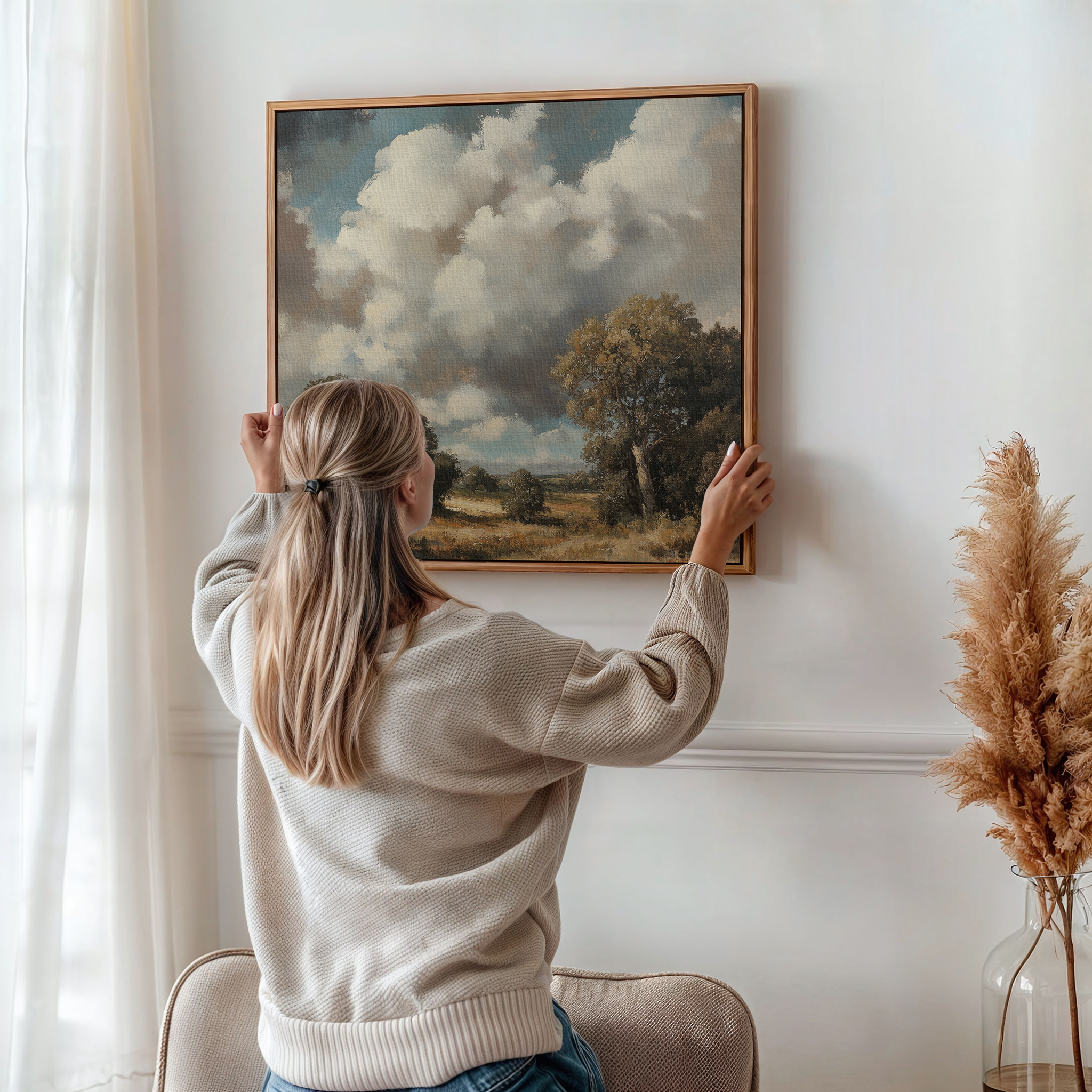 A woman with blonde hair in a beige sweater hangs the Gathering Storm Canvas Art—a framed landscape of clouds, trees, and grass—on a white wall in a softly lit room with natural decor.