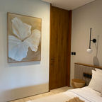 A minimal, modern bedroom features Aqua Flora Abstract Canvas Art above a wooden door, with a bedside table, a white hanging lamp, and part of an unmade bed with crisp white bedding visible.
