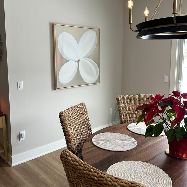 A dining area features a wooden table, four woven chairs, round placemats, and a poinsettia. The beige wall displays the Clover Harmony Abstract Canvas Art beneath a chandelier, brightened by natural light from the window.