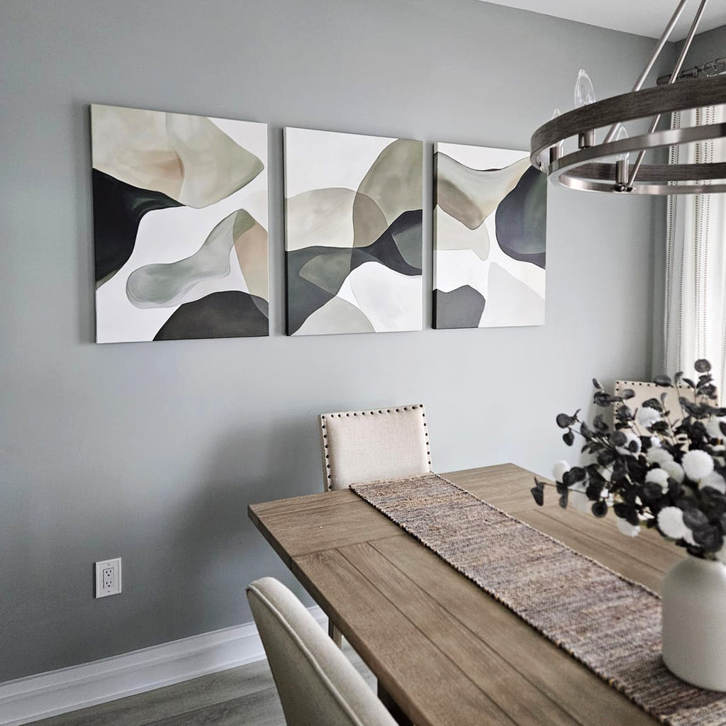 A modern dining room features a wooden table, beige upholstered chairs, and a vase with white and dark foliage. The Organic Flow Set of 3 abstract art in muted tones adds minimalist elegance to the gray wall.