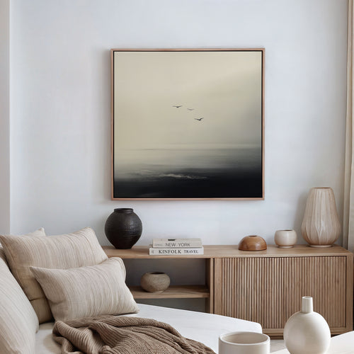 A minimalistic living room with neutral tones, a sofa with beige pillows, a wooden sideboard with vases, and the Above the Calm Canvas Art—minimalist wall art of three birds flying over a misty sea—perfect for large living room decor.