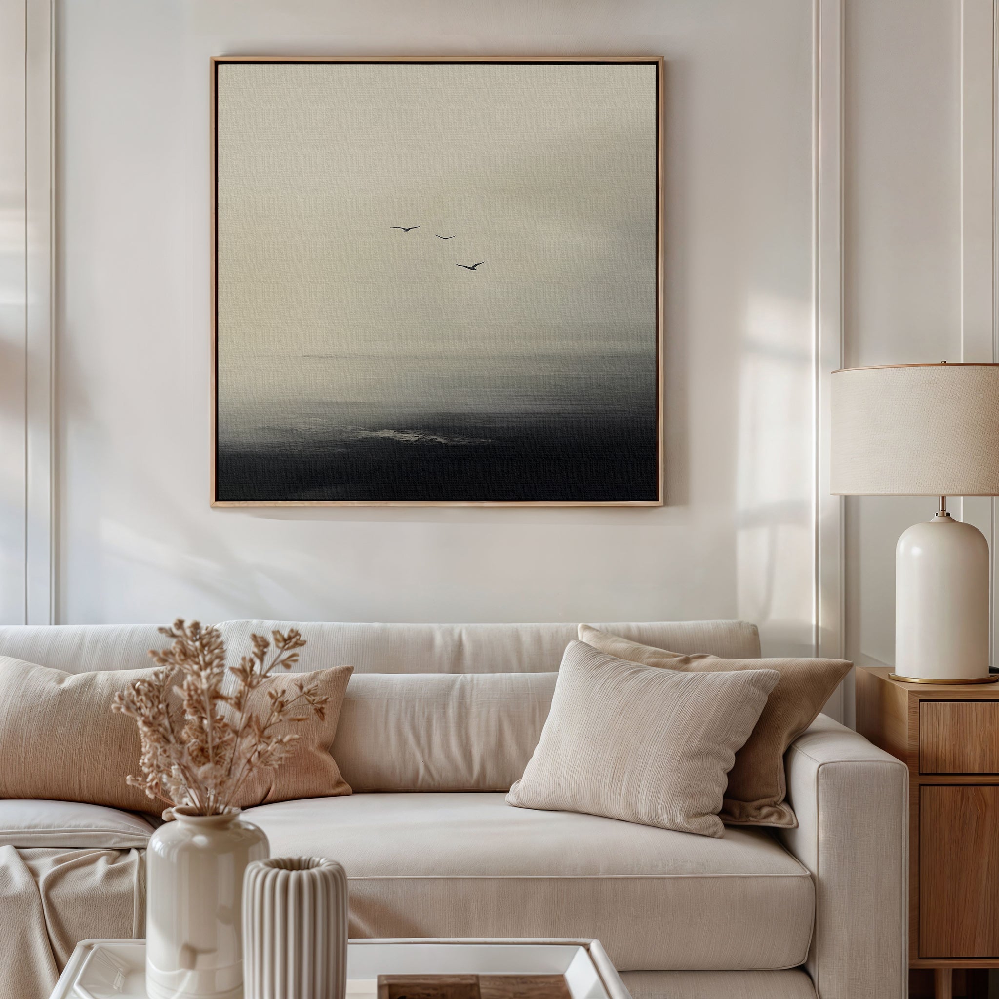 A modern living room with a beige sofa, neutral cushions, a side table with a cream lamp, and minimalist wall art—Above the Calm Canvas Art featuring three birds flying over a misty sea—perfect for large living room decor.