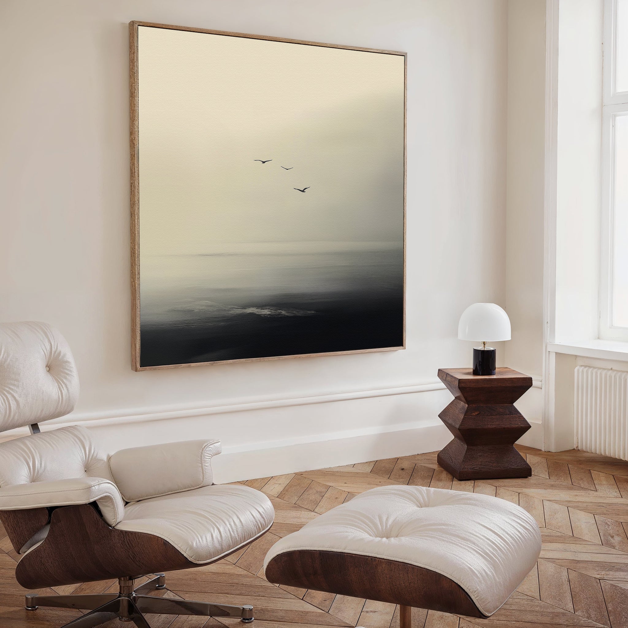 A modern living room with a white leather lounge chair and ottoman, wooden floors, a geometric wood side table with lamp, and minimalist Above the Calm Canvas Art featuring birds soaring over a misty landscape—ideal for spacious decor.