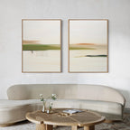 A modern living room features a curved beige sofa, a round wooden coffee table with two vases of flowers, and the Abstract Horizons Set of 2—minimalist abstract landscape paintings in wooden frames—hanging on a white wall.