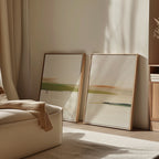 The Abstract Horizons Set of 2 features minimalist canvas art in light wood frames, leaning against a beige wall by an armchair. Soft sunlight highlights their earthy tones and modern style, creating a calming decor accent.