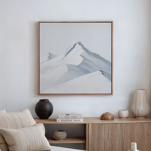 A minimalist living room features Alpine Ascent Abstract Canvas Art, neutral decor, a wooden sideboard, and vases by a stack of books including KINFOLK TRAVEL, creating a calm and modern atmosphere.
