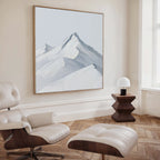 A modern living room with a white leather lounge chair and ottoman, wooden side table with a small white lamp, and the Alpine Ascent Abstract Canvas Art of snowy mountains completes the perfect decor.