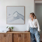 A woman in a white blouse and jeans leans against a wooden cabinet, admiring the Alpine Ascent Abstract Canvas Art of snowy mountains, which brings a serene touch to her living room decor. A small plant and books sit on the cabinet below.