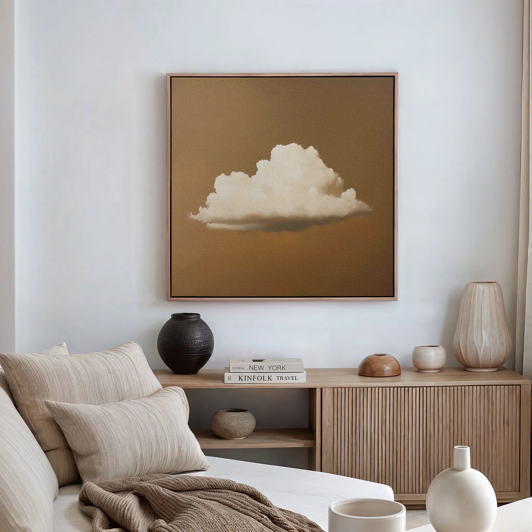 A minimalist living room features a beige couch with striped cushions, a light wood sideboard with vases and books, and modern wall art—Amber Sky Cloud Canvas Art, a framed painting of a white cloud on a brown background—above.