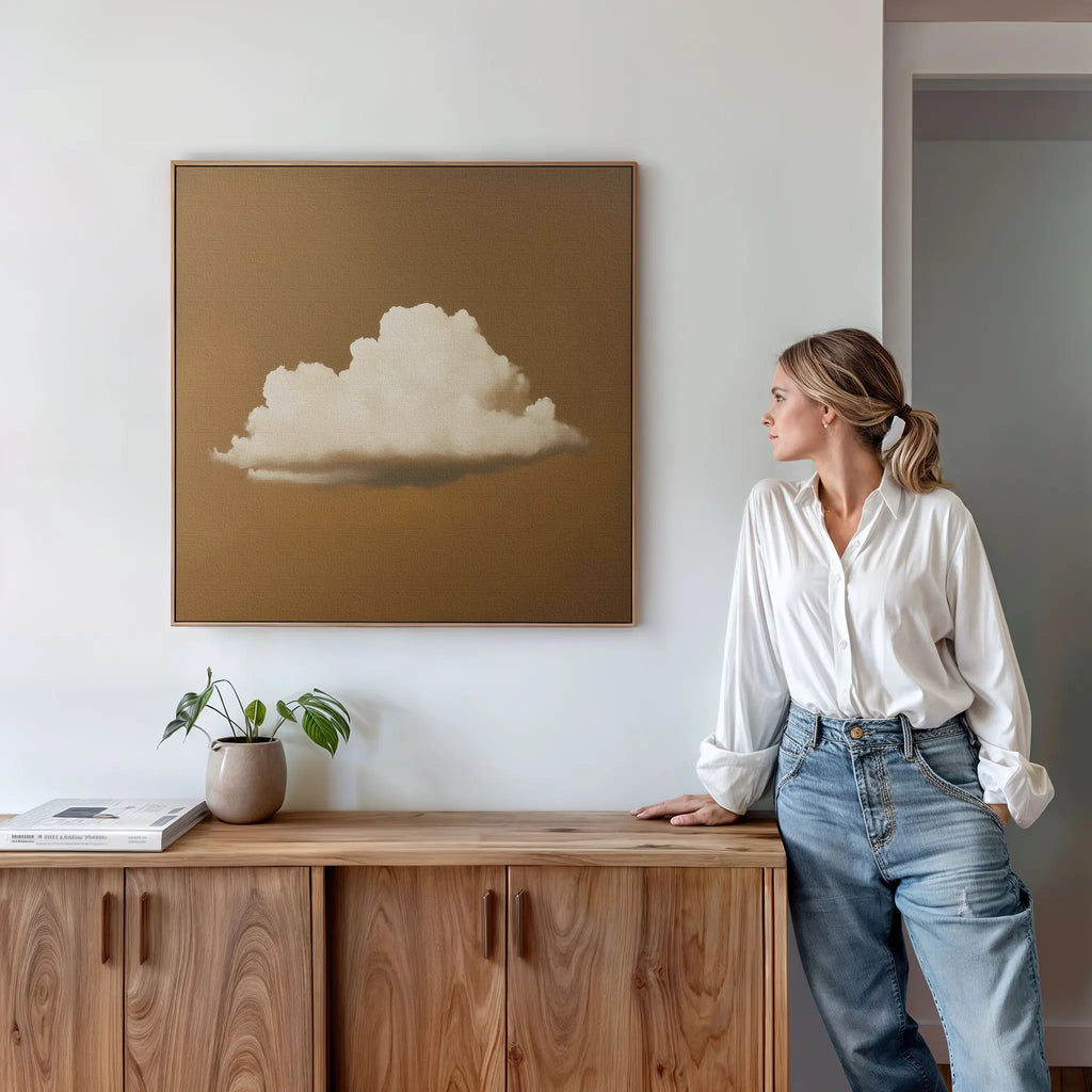A woman in a white blouse and jeans leans against a wooden sideboard, admiring the Amber Sky Cloud Canvas Art—a minimalist cloud painting on brown—hanging on a white wall. A potted plant and books complete the stylish bedroom decor.
