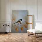 The Autumn Branch Canvas Art, featuring a chickadee on an autumn branch, leans against a white paneled wall in a room with wood flooring, a cushioned chair, and a round table holding candles and books.