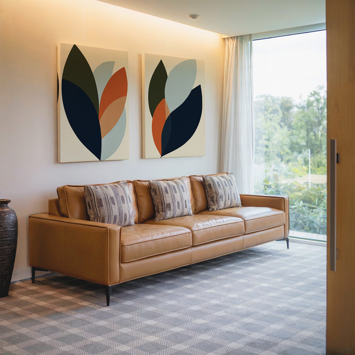 A modern living room featuring a tan leather sofa with Autumn Layers Set of 2 patterned pillows, abstract wall art, a large window with sheer curtains, a patterned carpet, and a decorative vase beside the sofa.