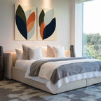 A modern bedroom features a large bed with white and gray bedding, decorative pillows, abstract Autumn Layers Set of 2 wall art above the headboard, tall vases on each side, and a big window overlooking greenery.