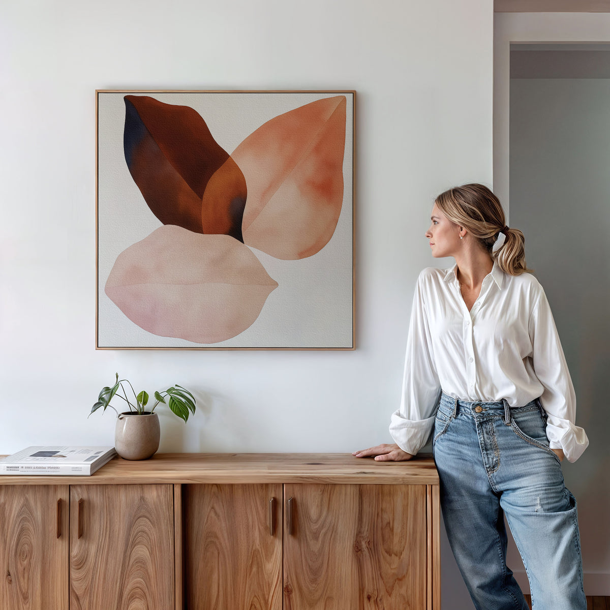 A woman in a white blouse and jeans leans against a wooden cabinet, gazing at the Autumn Petals Canvas Art with earthy tones above her. A small potted plant and a book sit on the cabinet.