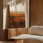 A soft, sunlit room features a wooden cabinet with the Autumn Plains Abstract Canvas Art propped against the wall, a round white table, and a textured white armchair, creating a warm, minimalist atmosphere.