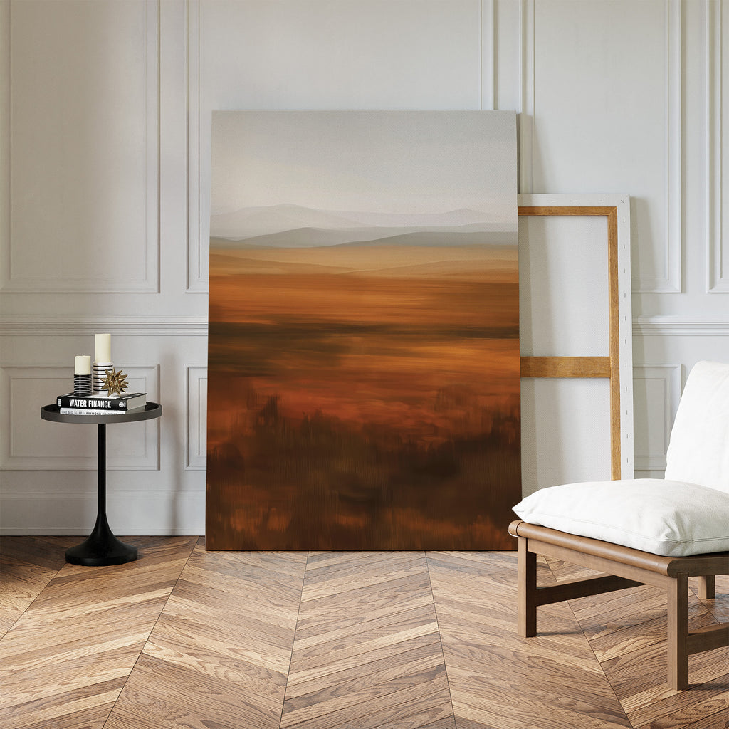 The Autumn Plains Abstract Canvas Art, with warm earth tones, leans against a white-paneled wall beside a wooden chair with white cushion and a round table holding candles and books on a chevron wood floor.