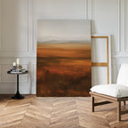 The Autumn Plains Abstract Canvas Art, with warm earth tones, leans against a white-paneled wall beside a wooden chair with white cushion and a round table holding candles and books on a chevron wood floor.