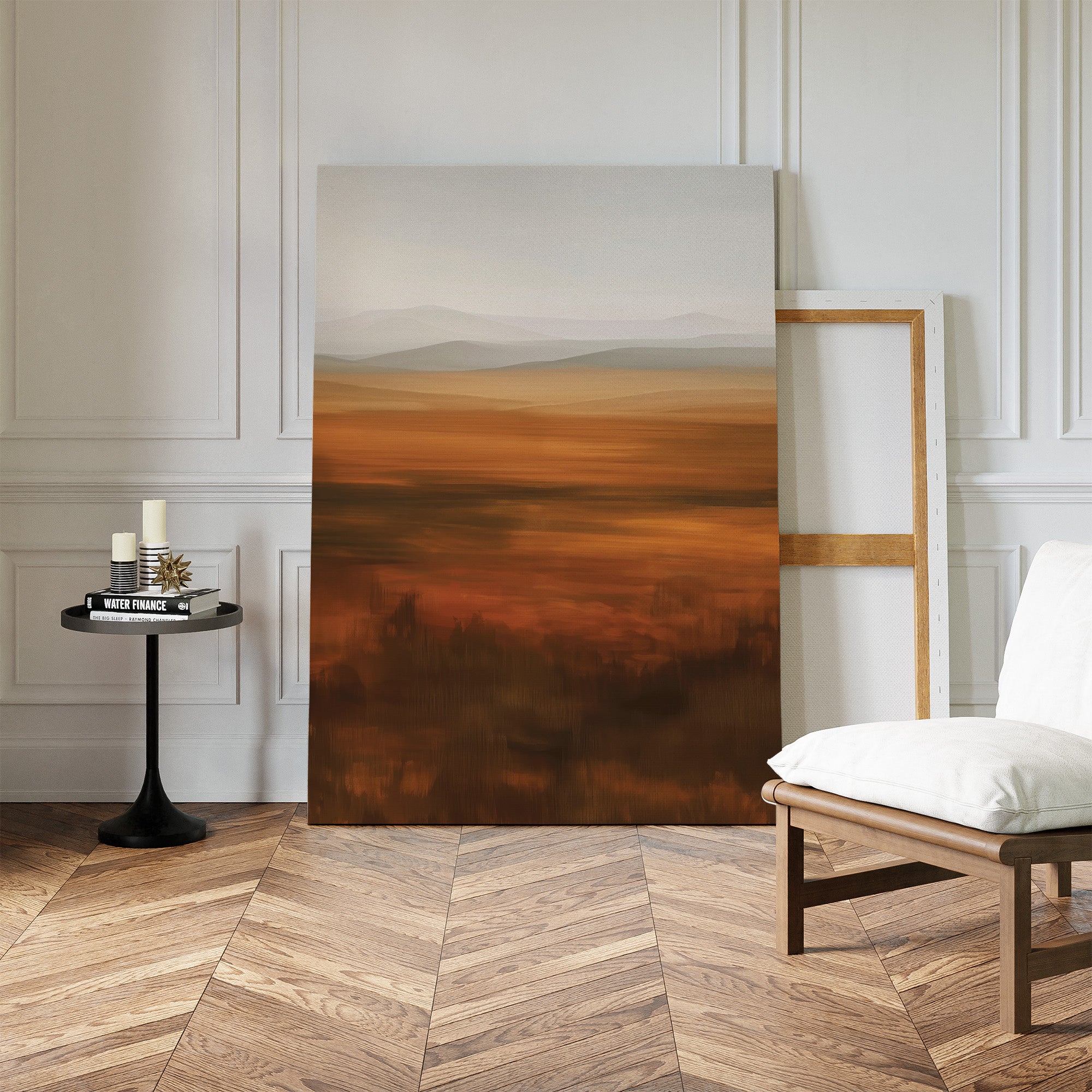 The Autumn Plains Abstract Canvas Art, with warm earth tones, leans against a white-paneled wall beside a wooden chair with white cushion and a round table holding candles and books on a chevron wood floor.
