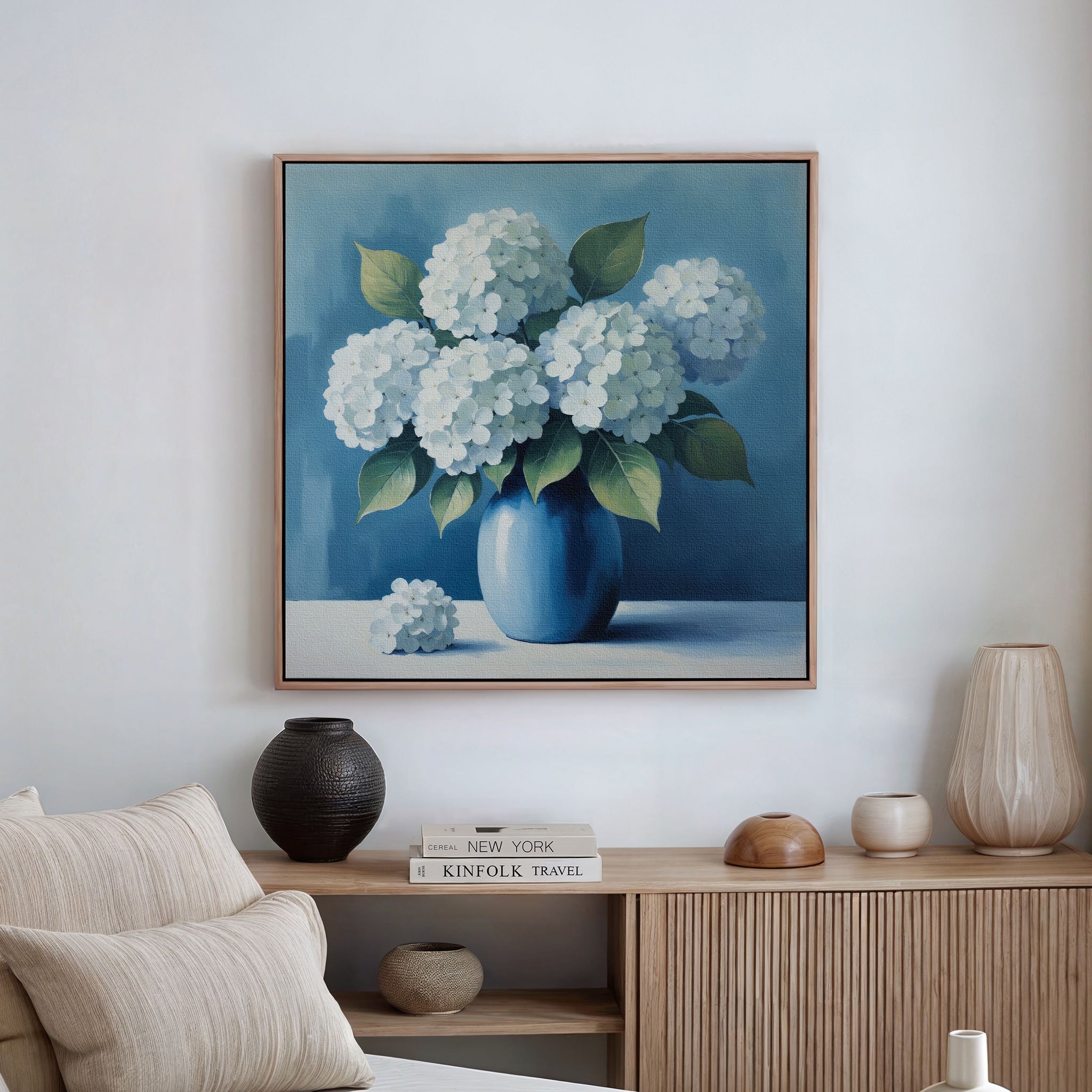 The Azure Cluster Canvas Art, a framed blue hydrangea print, hangs above a wooden sideboard with neutral pottery and decor—ideal floral wall art to enhance cozy living room decor. A light-colored sofa appears partially in the foreground.