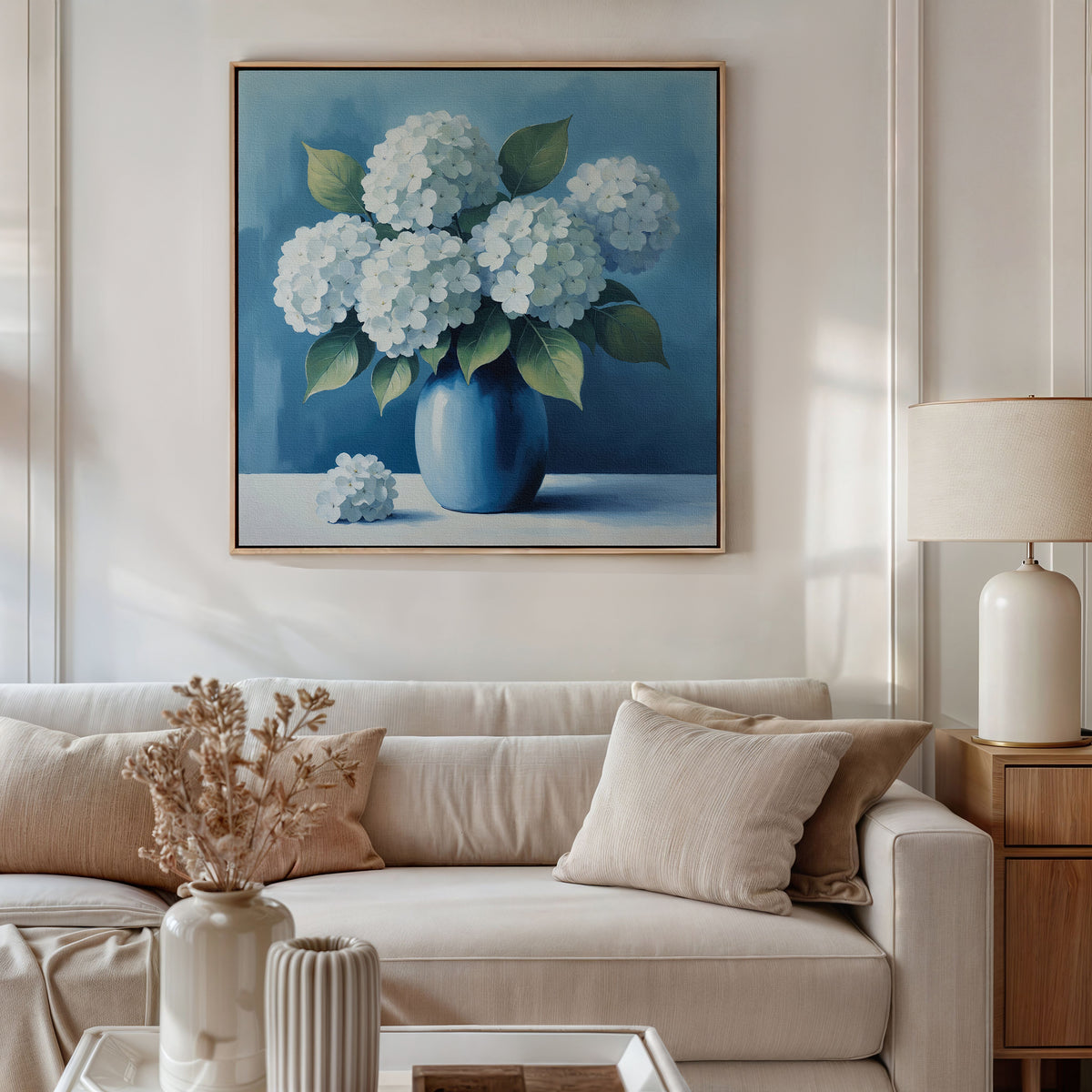 A cozy living room features a beige sofa, neutral cushions, side table with lamp, dried flowers in a vase, and the Azure Cluster Canvas Art with white hydrangeas in a blue vase above the sofa—ideal for elegant decor.