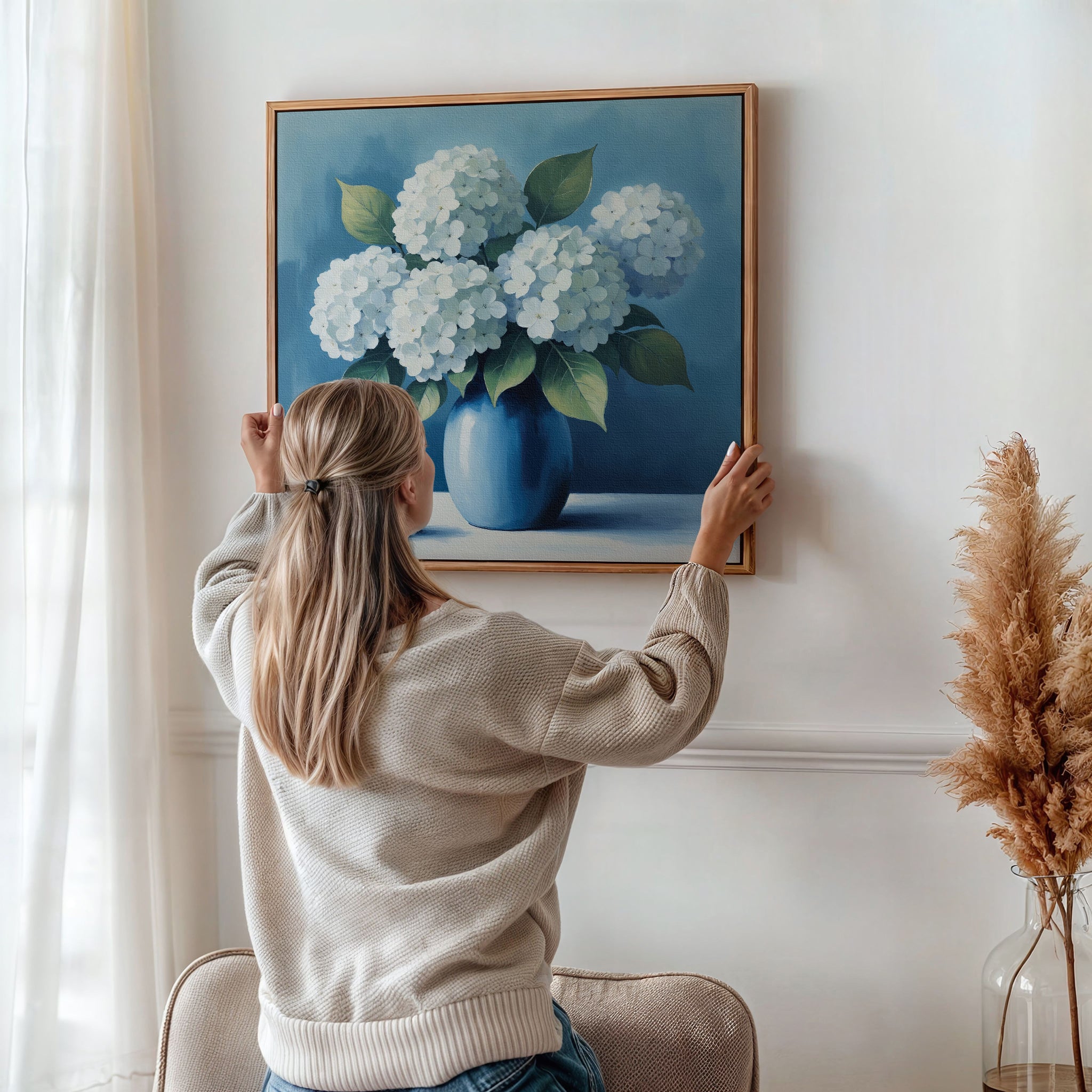 A woman with long blonde hair hangs the Azure Cluster Canvas Art on a white wall. Sunlight streams through a window, while dried pampas grass in a vase completes this elegant floral wall art for chic living room decor.