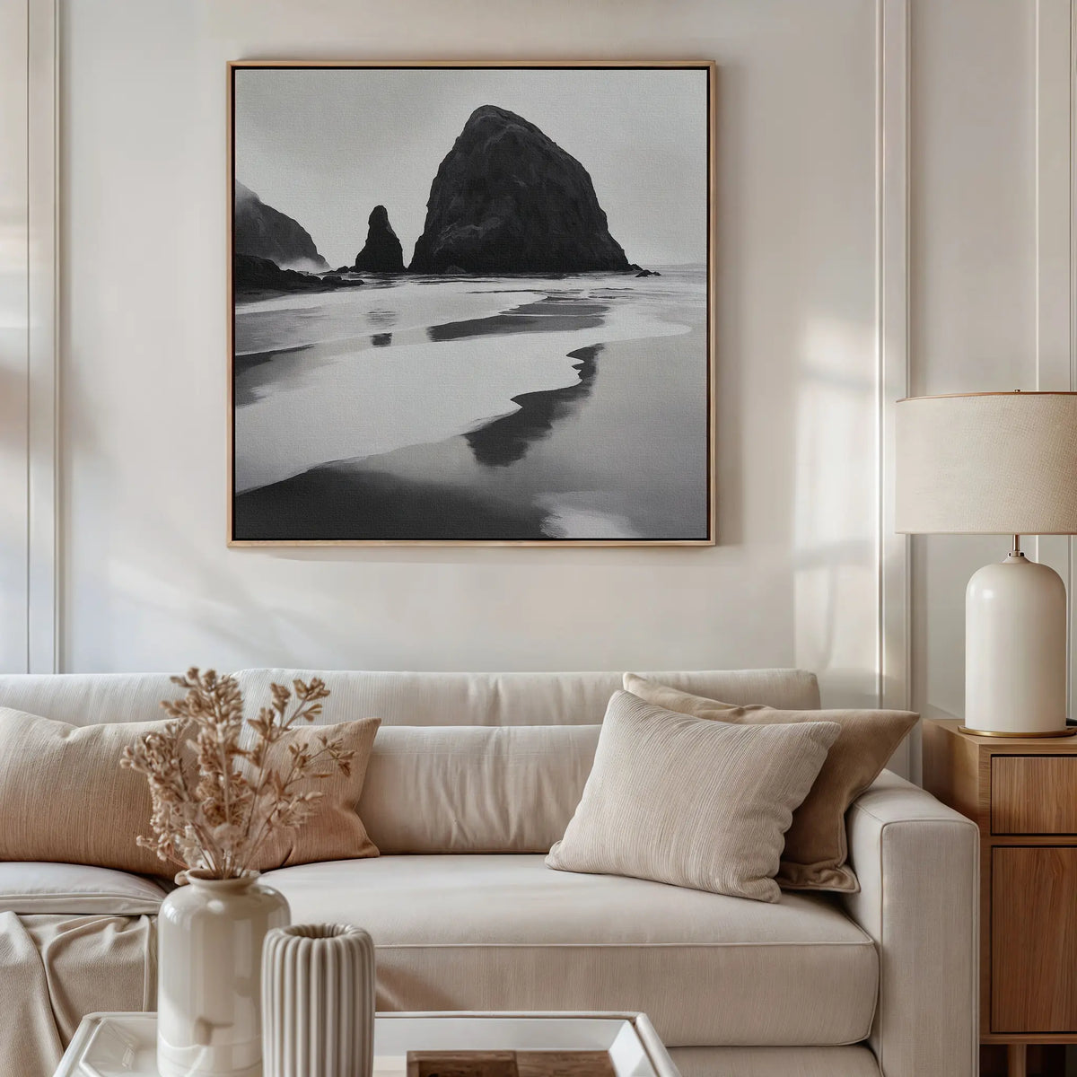 A neutral-toned living room features a beige sofa, cushions, a wooden side table with a white lamp, dried flowers in a vase, and the Basalt Beach Canvas Art above the sofa for a minimalist coastal look.
