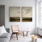 A modern, minimal living room with a gray sofa, wooden chair, small white side table, and three lit candles. Above the chair, the Beneath The Clouds Set of 2 canvas hangs on a white wall. Large windows fill the space with natural light.