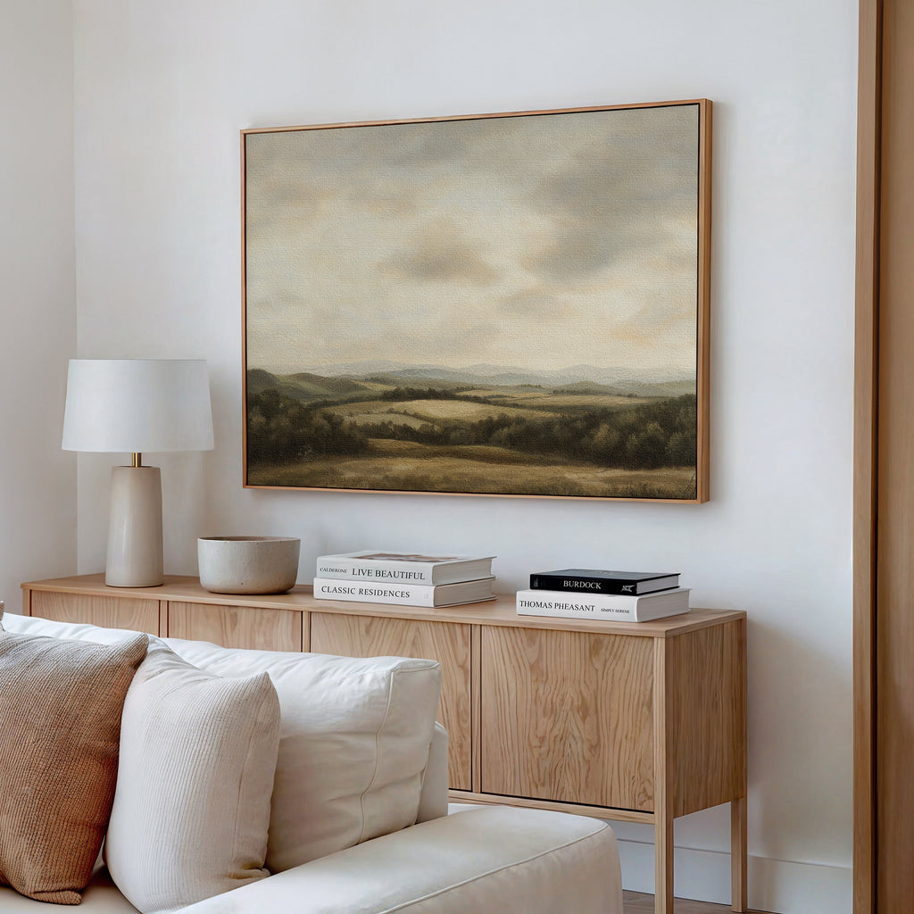 A modern living room displays a wooden sideboard with books, a lamp, and decor. Above it hangs the Beneath The Clouds Canvas Art, showcasing rolling hills under a cloudy sky. A white couch sits in the foreground.