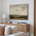 A modern living room displays a wooden sideboard with books, a lamp, and decor. Above it hangs the Beneath The Clouds Canvas Art, showcasing rolling hills under a cloudy sky. A white couch sits in the foreground.