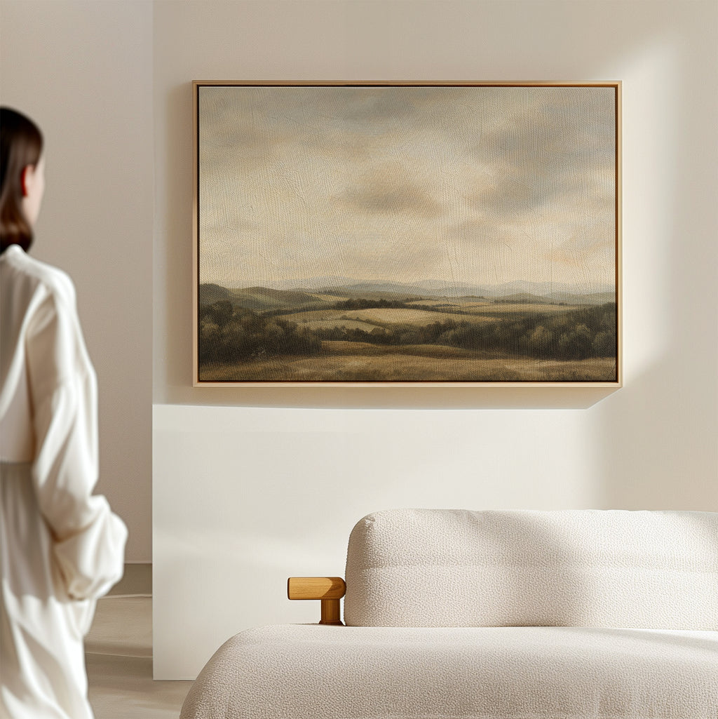 A person stands in a softly lit room, admiring Beneath The Clouds Canvas Art—a framed landscape of rolling hills and a cloudy sky—displayed above a light-colored sofa.