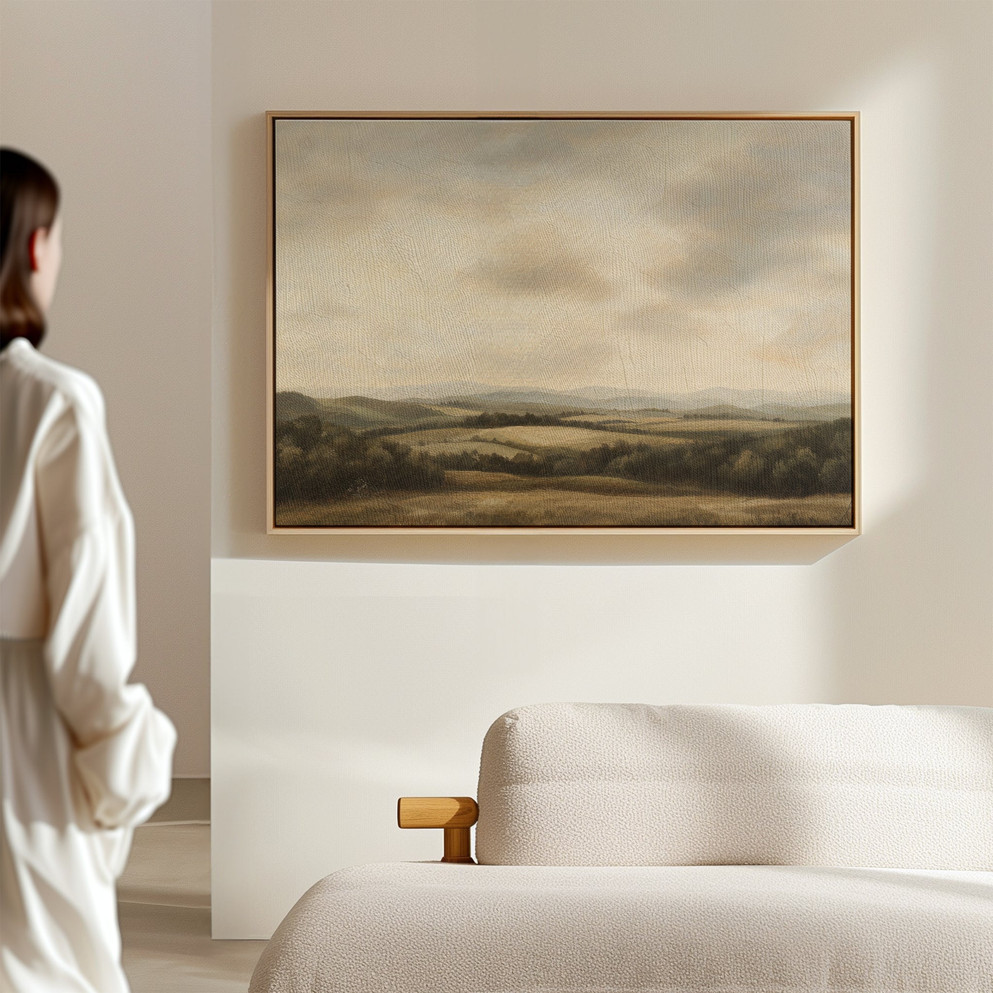A person stands in a softly lit room, admiring Beneath The Clouds Canvas Art—a framed landscape of rolling hills and a cloudy sky—displayed above a light-colored sofa.