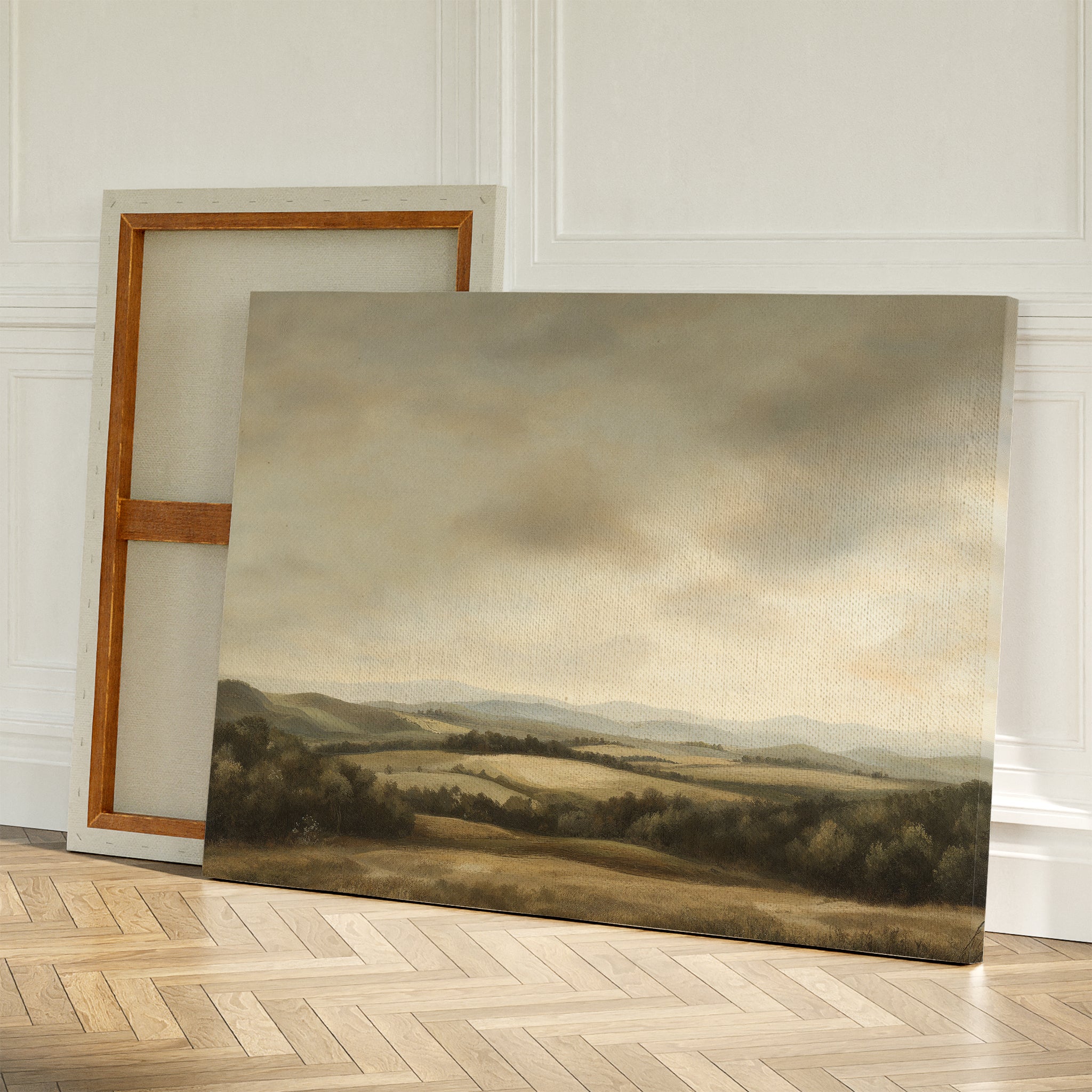 Two Beneath The Clouds Canvas Art pieces lean on a wooden floor against a white wall—one shows serene hills under cloudy skies while the other reveals its minimalist wooden frame.