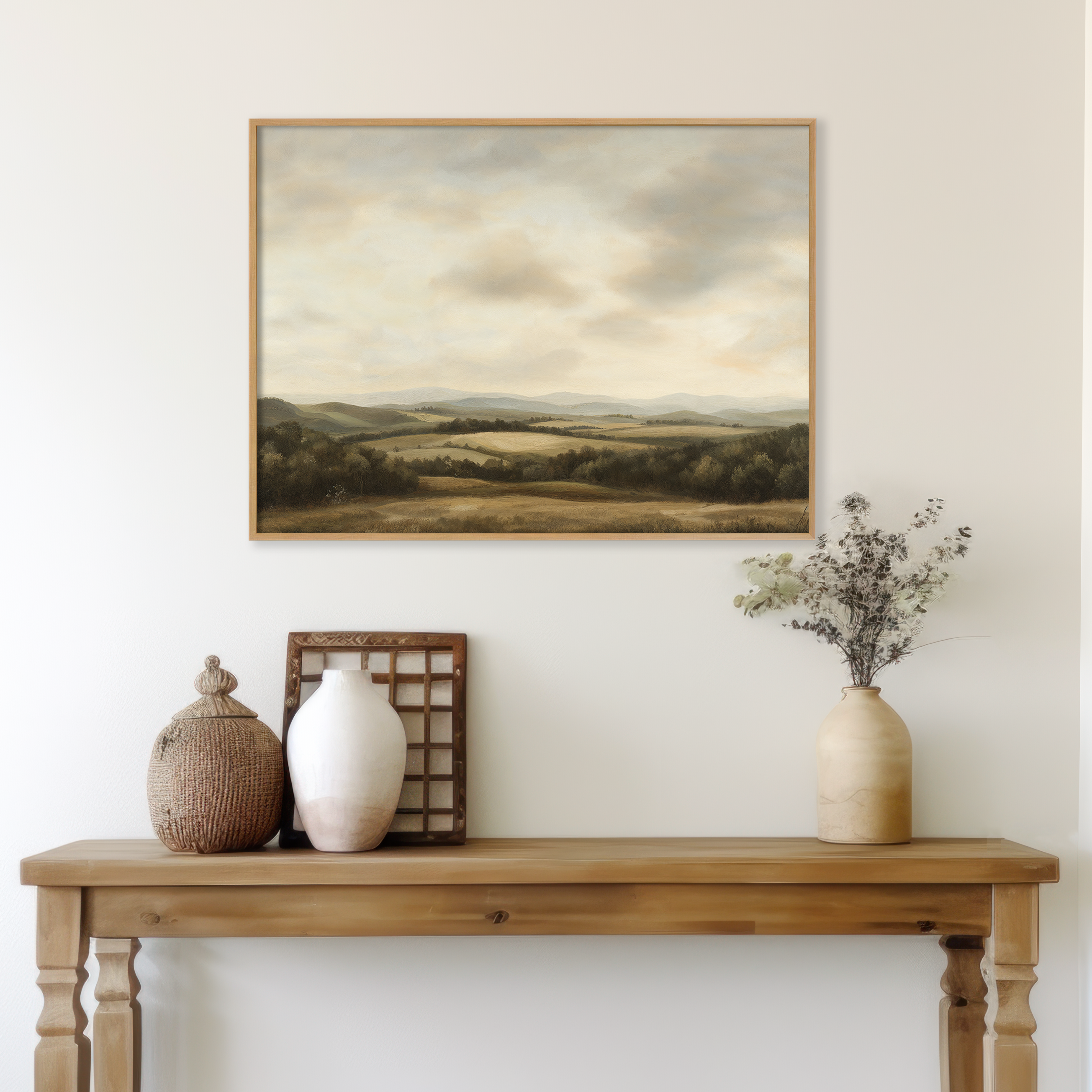 A wooden table with neutral-toned vases, a basket, and dried flowers sits against a white wall. Above the table hangs Beneath The Clouds Canvas Art, featuring a minimalist landscape of rolling hills under a cloudy sky.