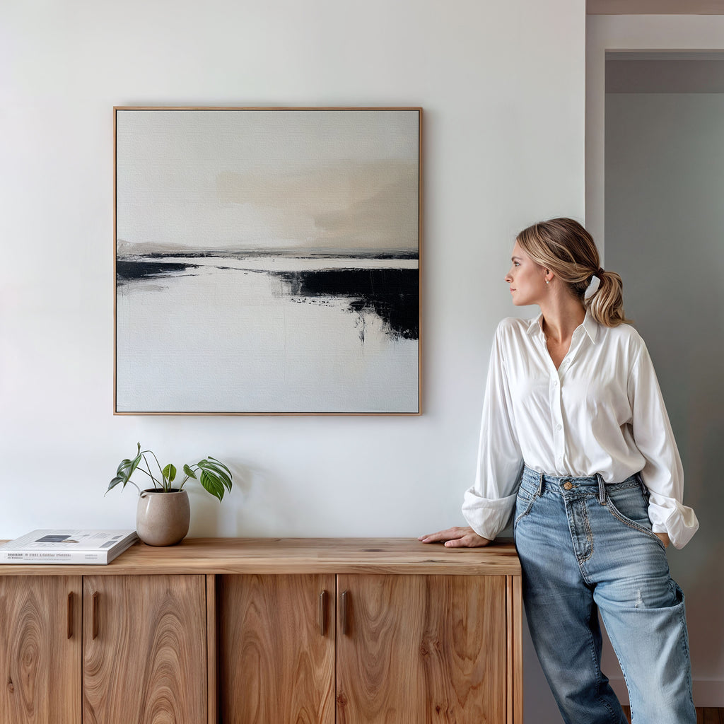 A woman in a white blouse and jeans leans on a wooden cabinet, admiring the Binary Calm Abstract Canvas Art in neutral tones. A small plant and a book on the cabinet enhance the minimal, modern elegance of the scene.