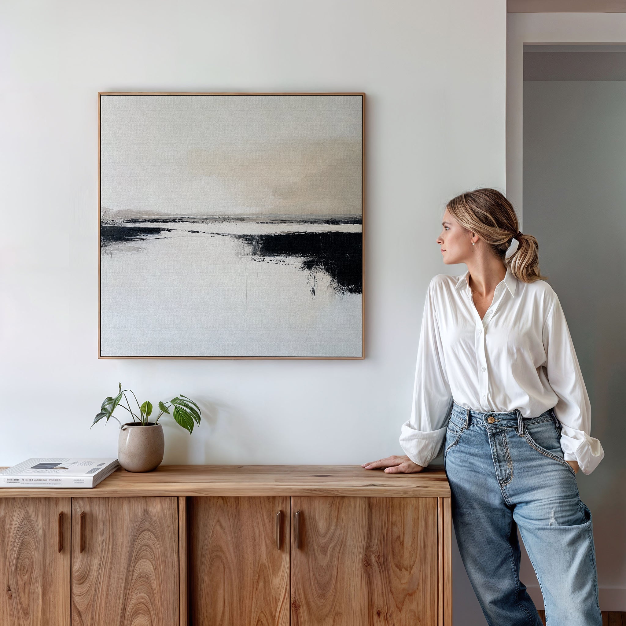 A woman in a white blouse and jeans leans on a wooden cabinet, admiring the Binary Calm Abstract Canvas Art in neutral tones. A small plant and a book on the cabinet enhance the minimal, modern elegance of the scene.
