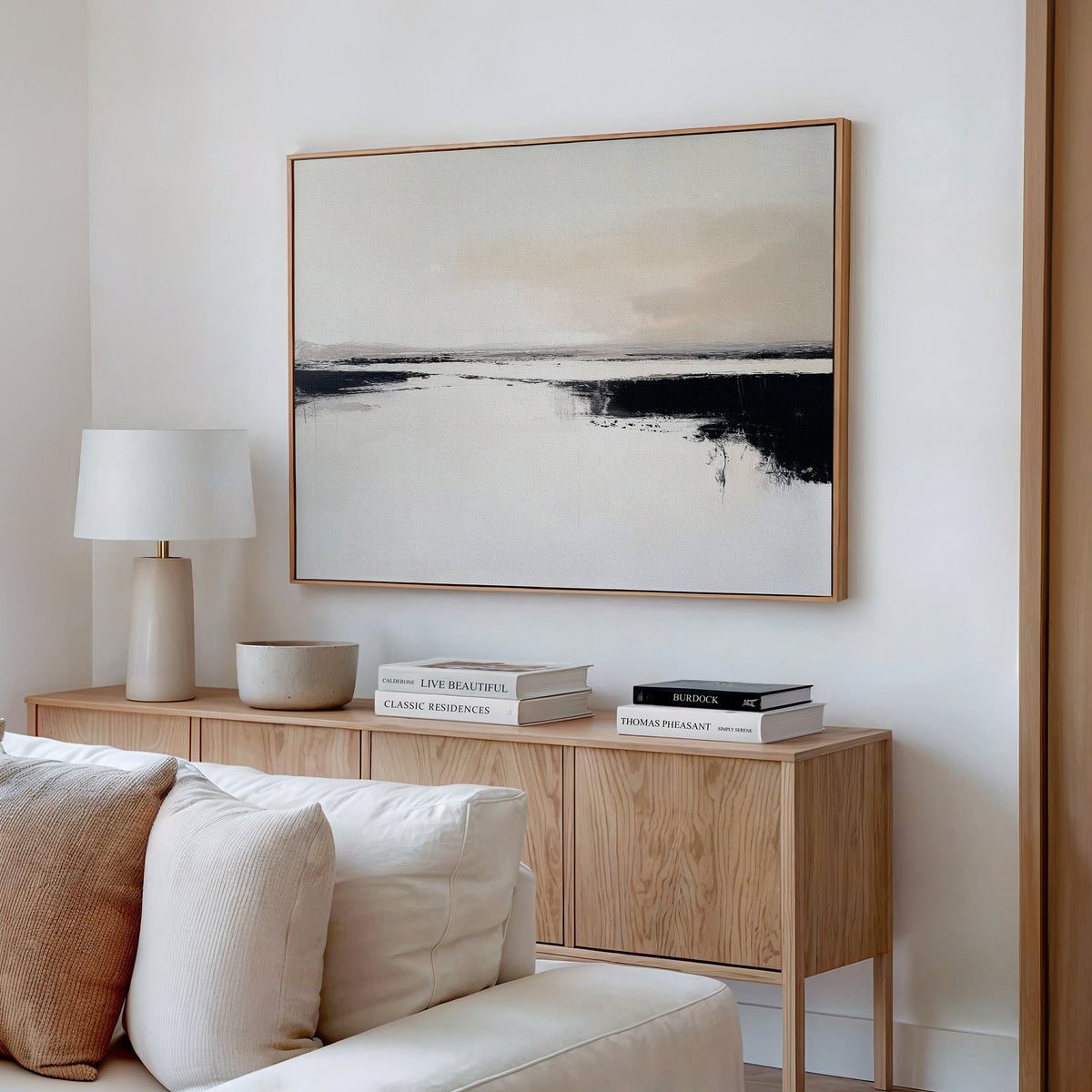 A minimal, modern living room features a wooden sideboard topped with stacked books, a lamp, and a bowl. Above it hangs the Binary Calm Abstract Canvas Art – Horizontal in neutral tones. A white sofa with cushions sits in the foreground.