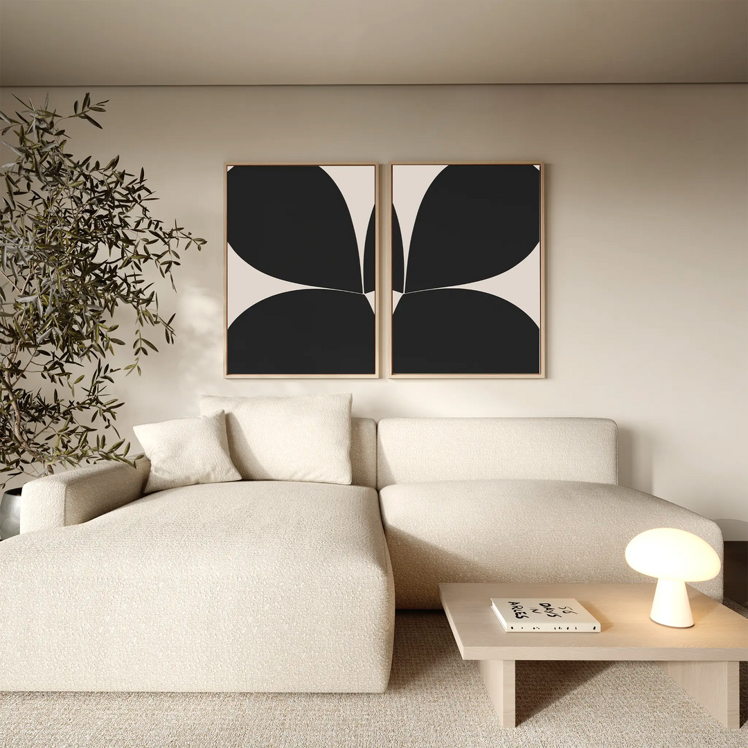 A modern living room with a cream sectional sofa, light wood coffee table, indoor plant, and the Minimal Petal Set of 2—a pair of minimalist black-and-white abstract wall art pieces—displayed above the seating area.