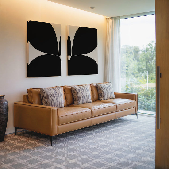 A modern living room with a tan leather sofa, patterned pillows, minimalist Minimal Petal Set of 2 wall art, a large window with sheer curtains, and a decorative vase on the floor.