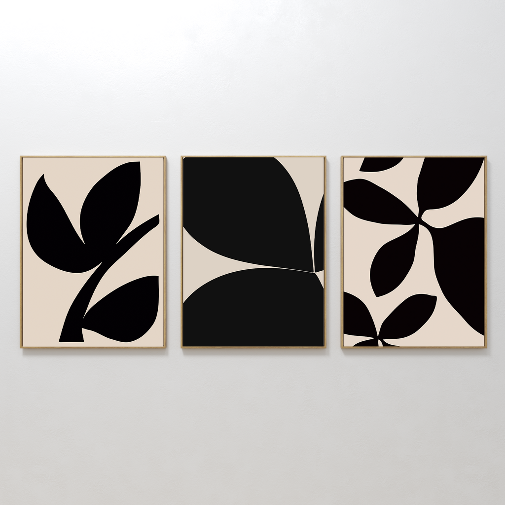 The Black Botanical Set of 3 features minimalist art prints with abstract black leaf designs on beige backgrounds, each framed in light wood and displayed side by side for striking botanical wall art.