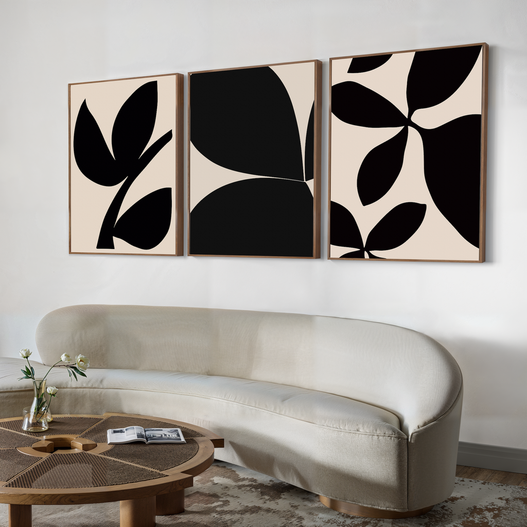 A minimalist living room features a curved beige sofa, a round wooden coffee table with decor, and the Black Botanical Set of 3—Nordic park-inspired botanical wall art—displayed above.