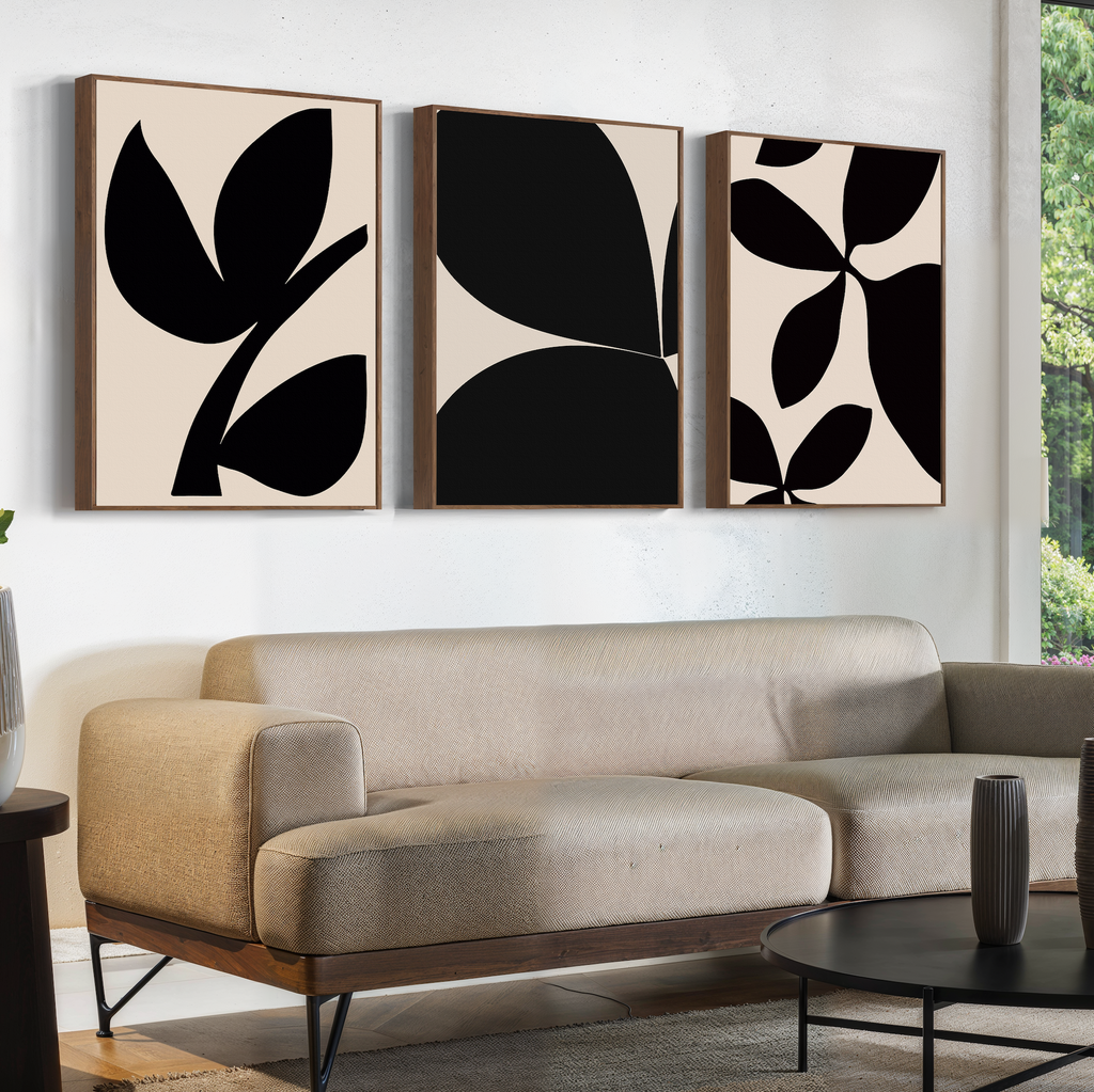 A modern living room features a beige sofa, a round black coffee table, and the Black Botanical Set of 3 abstract black-and-white prints hanging above the sofa. Natural light streams in from a window on the right.