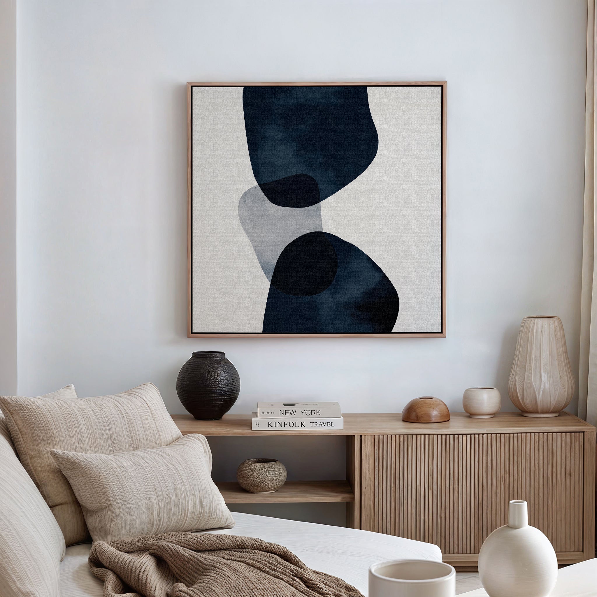 A minimalist living room with a beige sofa, striped cushions, a wooden cabinet decorated with vases and books, and striking modern wall art—Blue Currents Canvas Art—featuring dynamic organic shapes in black and gray.