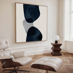 A modern living room featuring a white lounge chair and ottoman, geometric wooden side table with a white lamp, and Blue Currents Canvas Art on a cream wall above light wood herringbone flooring.