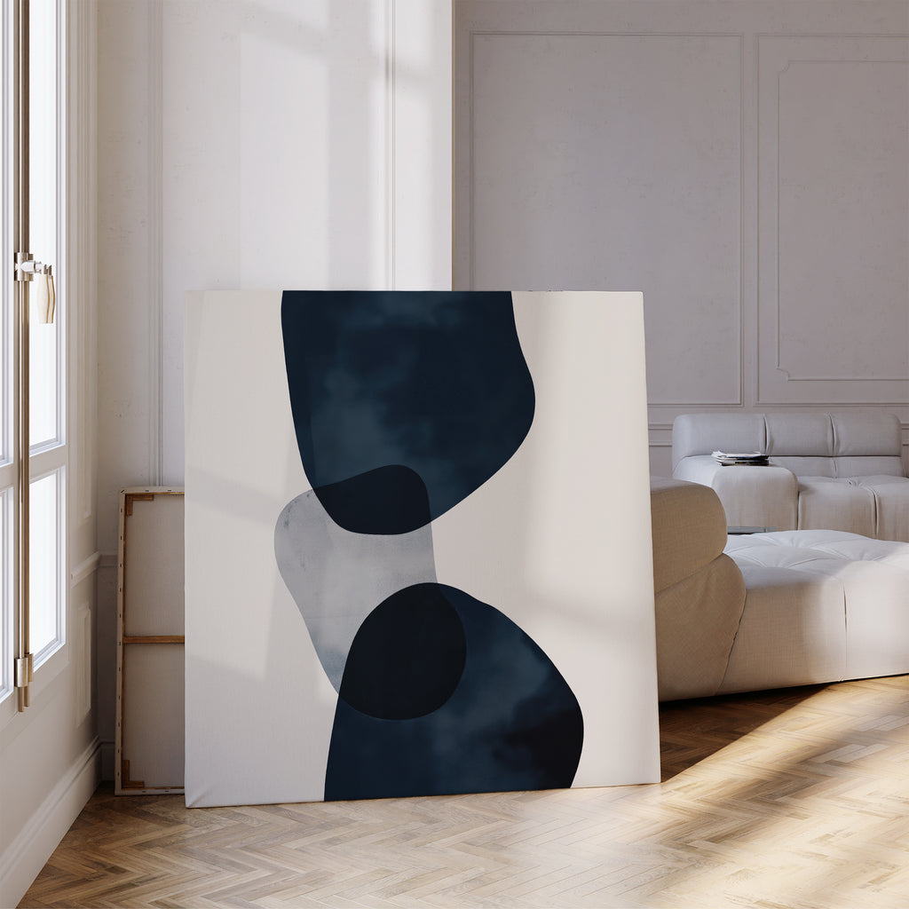 The Blue Currents Canvas Art, featuring abstract black and gray shapes, leans against a wall in a bright living room with a beige sofa and parquet flooring, as sunlight pours through tall windows.