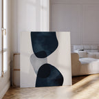 The Blue Currents Canvas Art, featuring abstract black and gray shapes, leans against a wall in a bright living room with a beige sofa and parquet flooring, as sunlight pours through tall windows.