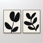 The Black Leaf Set of 2 features framed minimalist prints, each with striking black botanical silhouettes—three leaves on one, five on the other—set against light backgrounds. Perfect for fans of minimalist or botanical wall art.