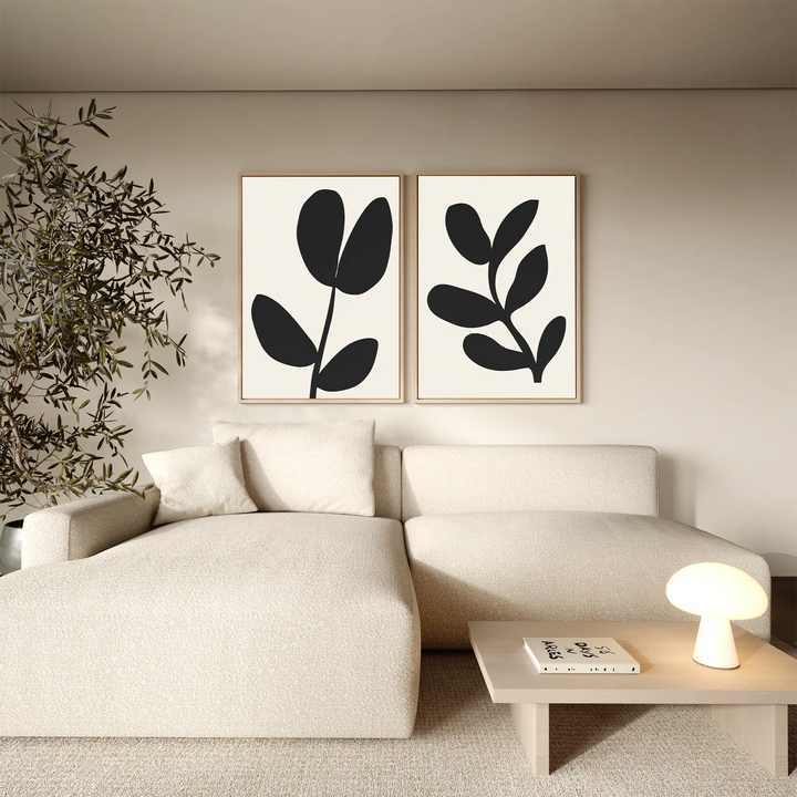 A minimalist living room featuring a light beige sectional sofa, wooden coffee table with lamp and book, potted plant, and the Black Leaf Set of 2 wall art prints for a modern botanical touch.